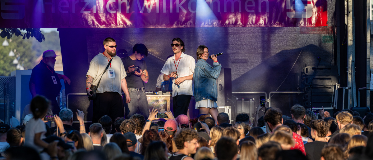 Eine Rap-Crew performt mit sichtbarer Energie auf der Bühne. Die Menschenmenge vor der Bühne tanzt mit, während lila Scheinwerfer die Szene beleuchten.