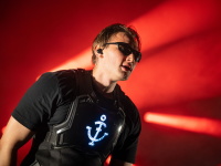 Künstler der Band trägt eine leuchtende Weste mit LED-Anker-Logo, Sonnenbrille und schaut in die Menge. Der Bühnenhintergrund ist in rotes Licht getaucht.
