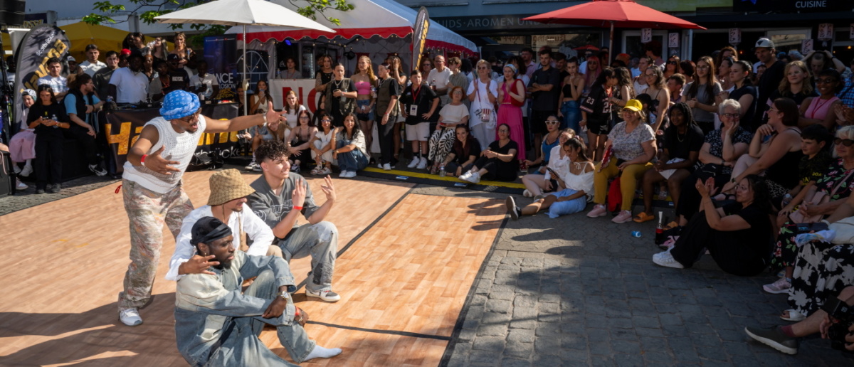 Vier Tänzer performen auf einem Platz vor Zuschauern, einer steht gestikulierend, drei sitzen.
