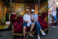 Zwei Personen sitzen lachend auf einem roten, barocken Sofa zwischen zwei goldfarbenen Theaterfiguren vor einer bunt gestalteten Festival-Kulisse.