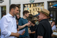 Zwei Männer unterhalten sich lächelnd vor einem Lokal, während im Hintergrund weitere Gäste zusammensitzen.