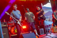 Musiker einer Rockband bewegen sich dynamisch über die Bühne, während rotes Licht und Bühnennebel die Szene prägen.