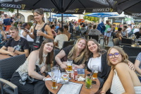 Eine Gruppe junger Frauen sitzt lächelnd an einem vollen Außentisch eines Lokals.
