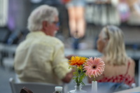 Zwei Blumen in kleinen Vasen stehen im Vordergrund, während sich zwei Personen unscharf im Hintergrund unterhalten.