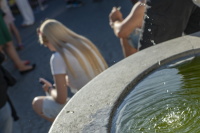 Ein Brunnenrand mit spritzendem Wasser steht im Vordergrund, während Personen im Hintergrund unscharf sitzen.