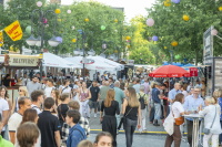 Eine breite Feststraße ist mit Essensständen, Sonnenschirmen, Lampions und vielen Besucherinnen und Besuchern gefüllt.