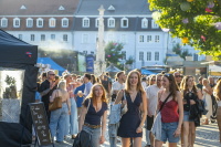 Drei junge Frauen laufen lächelnd über einen sonnigen Platz voller Festivalgäste.