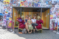 Eine Fotowand mit vielen bunten Altstadtfest-Plakaten rahmt ein rotes Sofa mit drei sitzenden Personen und zwei gold geschminkten Darstellerinnen.