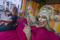 Eine gold geschminkte Darstellerin lehnt hinter einem roten Sofa und zeigt lachend auf eine Person am Bildrand.