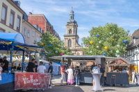 Ein sonniger Platz mit Markt- und Getränkeständen liegt vor einer Kirche und ist mit bunten Lampions geschmückt.