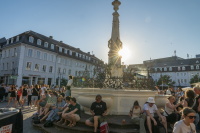 Viele Menschen sitzen rund um einen Brunnen auf einem sonnigen Platz.