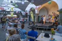 Publikum sitzt in einem Innenhof vor einer kleinen Bühne mit zwei Personen am Mikrofon.