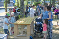 Kinder spielen an einer Holzstation unter Bäumen, daneben stehen Erwachsene und Kinderwagen.