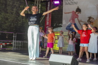 Kinder tanzen mit einer Anleiterin auf einer Bühne im Freien.