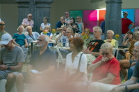 Publikum sitzt an Tischen in einem Innenhof und schaut zu einer Veranstaltung.