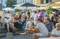 Menschen sitzen und stehen an Tischen auf einem belebten Platz in der Abendsonne.