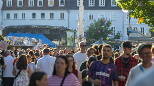 Viele Menschen drängen sich auf einem sonnigen Platz mit Brunnen und Marktständen.