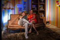 Zwei Personen sitzen auf einem Sofa in einer bunt gestalteten Kulisse.